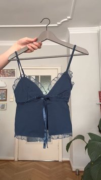 Eine Hand hält einen Holzkleiderbügel mit einem navyblauen ärmellosen Oberteil, das mit Rüschensaum und einem satinierten Band an der Front verziert ist, in einem hellen Raum mit gerahmten Fotos.
