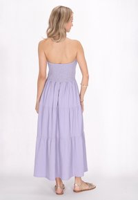 Maxikleid in Helllila, mit gerafftem Oberteil, gestuftem Design, strukturiertem Stoff, fließendem Rock und freiem Rücken. Kombiniert mit flachen Sandalen.