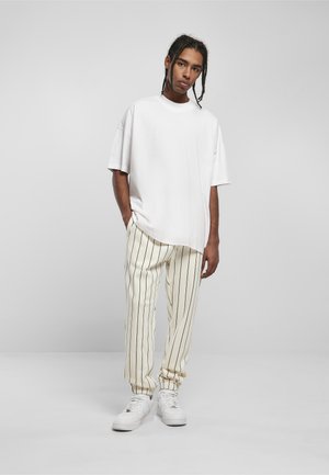 Young man standing with hands in pockets wearing oversized white T-shirt, cream pants with vertical black stripes, and white sneakers.