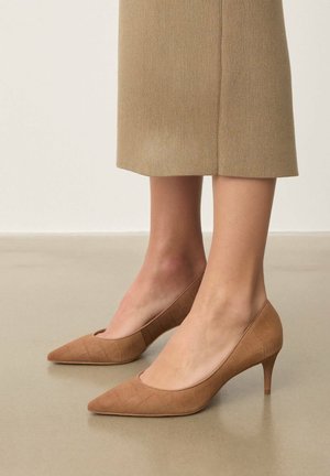 Világosbarna hegyes orrú magassarkú cipő texturált négyzetmintával, alacsony tűsarkú, puha anyagból. Semleges színű bordázott ruhával viselve.