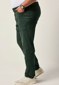 Uomo in pantaloni verde scuro con polsini elastici, sneakers bianche e un orologio da polso nero, in piedi su uno sfondo chiaro e uniforme.