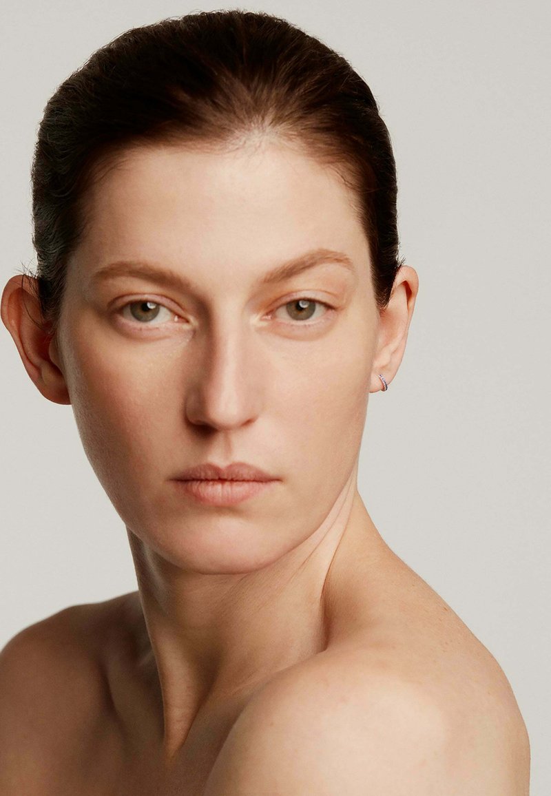 Primer plano de una persona con piel clara, cabello castaño recogido, que lleva un pequeño aro en la oreja, fondo neutro y maquillaje mínimo.