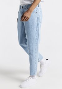 Jeans de mezclilla azul claro con un corte relajado, que cuentan con cinco bolsillos y cierre de botón. Combinados con zapatillas blancas y calcetines grises.
