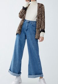 Cardigan à imprimé léopard porté sur un haut blanc à col montant, associé à un jean bleu à jambes larges avec revers et des baskets blanches.