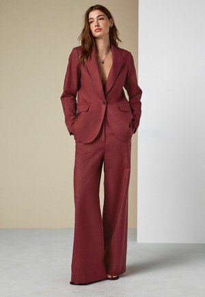 Frau steht mit den Händen in den Taschen, trägt einen dunkelroten Hosenanzug mit weitem Bein und offenem Blazer vor einem beige-weißen Hintergrund.