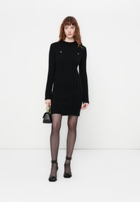 Abito nero a maglia con maniche lunghe, bottoni sul colletto e orlo a coste. Indossato con collant trasparenti e sandali neri con cinturini.