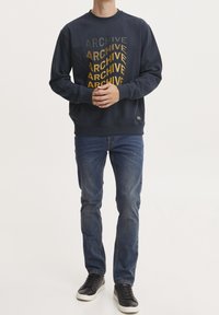 Sudadera marina con "ARCHIVE" impreso en amarillo y gris. Cuello redondo, mangas largas, combinada con jeans azules y sneakers negros.