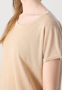 Kvinde med langt lyst brunt hår iført beige kortærmet striktrøje, vist fra nakke til overarm mod ensfarvet baggrund.