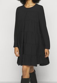 Robe noire à manches longues avec couches superposées, encolure ronde, coupe décontractée et texture fluide. Portée avec des bottes noires montant jusqu'aux genoux.