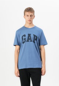 Ung mand med kort hår iført en blå GAP t-shirt og sorte bukser, stående foran en ensfarvet hvid baggrund.