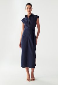 Abito camicia in lino navy con maniche corte strutturate, parte anteriore con bottoni, cintura in vita e spacchi laterali. Abbinato a sandali con tacco nudo.