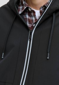 Veste noire zippée avec capuche, dotée d'une fermeture éclair gris clair et de cordons de serrage réglables. Portée par-dessus une chemise à carreaux rouge et blanc.