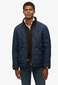 Man in een marineblauwe gewatteerde jas over een bruine ritssweater en een beige shirt, met blauwe jeans en een horloge, staand tegen een witte achtergrond.