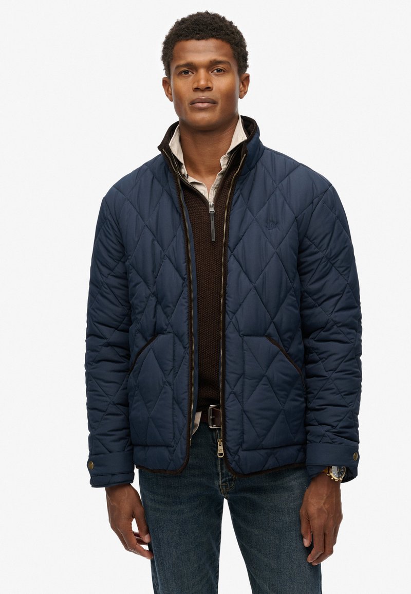 Man in een marineblauwe gewatteerde jas over een bruine ritssweater en een beige shirt, met blauwe jeans en een horloge, staand tegen een witte achtergrond.