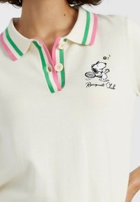 Witte polo met een roze en groene gestreepte kraag, met een Snoopy tennisafbeelding en de tekst "Racquet Club". Twee knoopsluiting.