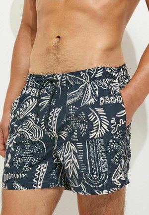 Męski tors w granatowych kąpielówkach z białymi abstrakcyjnymi wzorami plemiennymi, ręce w kieszeniach, stojący na jednolitym tle.