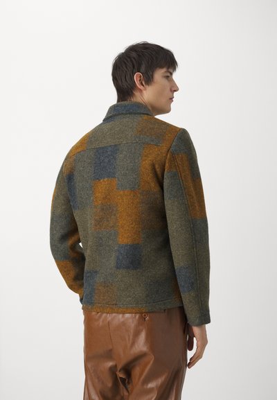 Wool jacket with a patchwork design in olive green, rust, and blue shades. Features a collar and straight-cut sleeves, paired with brown pants.
