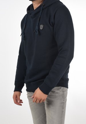 Mann trägt einen dunkelblauen Kapuzenpullover mit Kordeln und hellgraue Jeans, steht vor einem schlichten weißen Hintergrund.