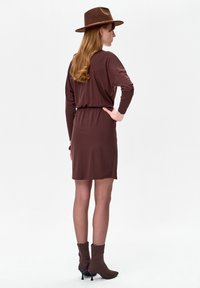 Kobieta stojąca tyłem, ubrana w brązową sukienkę z długim rękawem, brązowe botki na obcasie oraz szerokokapelowy brązowy kapelusz na białym tle.