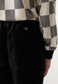 Personne portant un pantalon Vans noir avec une chemise à manches longues à carreaux noirs et blancs, main détendue le long du corps.