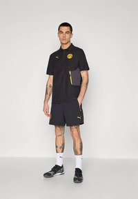 Čierne polo tričko so žltými detailmi a logom, v kombinácii s tmavými šortkami so zipsovými vreckami. Na nohách nosí čierne topánky a biele ponožky.