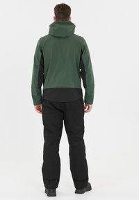 Veste imperméable verte avec une capuche et des accents noirs, associée à un pantalon noir. Elle présente un design élégant et des poignets ajustables.