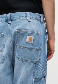 Osoba nosi svetlomodré riflové nohavice s veľkými zadnými vreckami, jedno vrecko má hnedý nášivku s logom Carhartt.