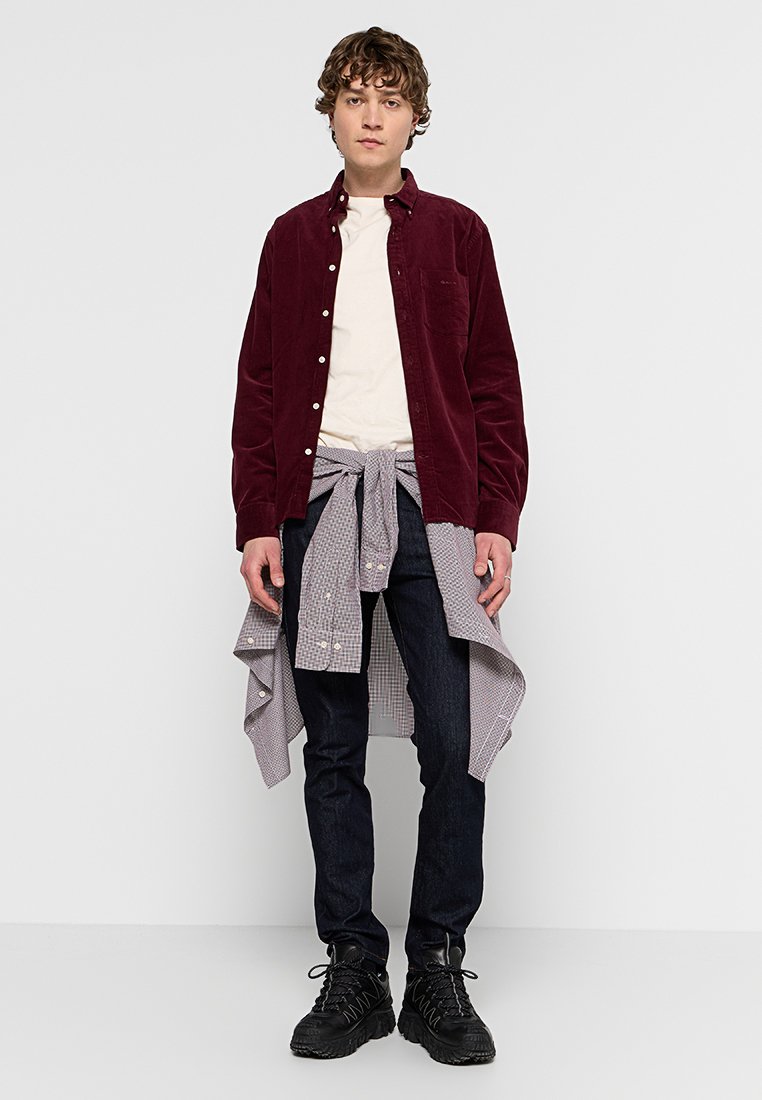 Bordeauxfarbendes Cordhemd, weißes T-Shirt, kariertes Hemd um die Taille gebunden, dunkle Jeans und schwarze Schnürstiefel mit strukturierten Sohlen.