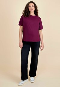 Maroon kortef mouwen t-shirt gecombineerd met zwarte wijde jeans en witte sneakers. Achtergrond is een lichte beige kleur.