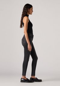 Top nero senza maniche con colletto alto, abbinato a jeans neri attillati. Le scarpe sono mocassini neri lucidi. Silhouette semplice e moderna.