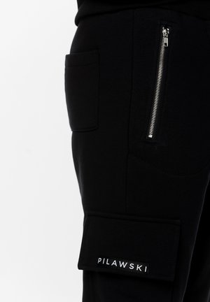 Czarne bawełniane spodnie cargo mają kieszeń na zamek i boczne kieszenie naszywane. Materiał jest miękki o gładkiej teksturze, zdobiony etykietą z logo.