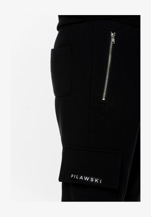 Czarne bawełniane spodnie cargo mają kieszeń na zamek i boczne kieszenie naszywane. Materiał jest miękki o gładkiej teksturze, zdobiony etykietą z logo.