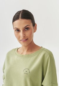 Felpa verde chiaro con scollatura ampia, texture morbida e grafica circolare blu "IL FUTURO VERDE INIZIA ORA" sul davanti.