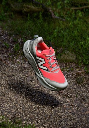 Chaussure de course New Balance corail vif et grise flottant au-dessus d'un sentier de gravier avec de l'herbe verte en arrière-plan.
