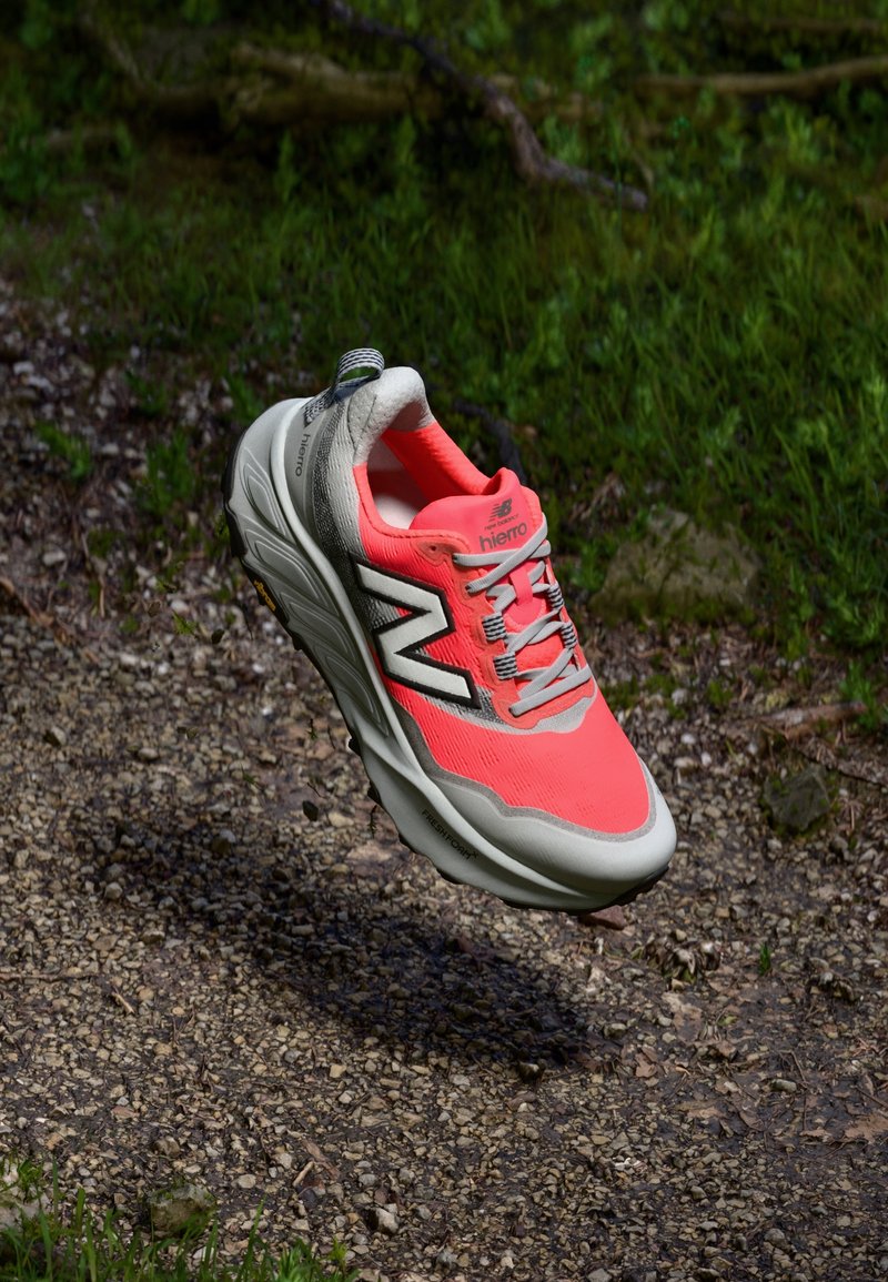 Chaussure de course New Balance corail vif et grise flottant au-dessus d'un sentier de gravier avec de l'herbe verte en arrière-plan.