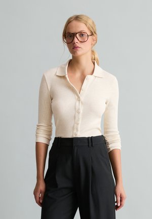 Frau mit blondem Haar zu einem niedrigen Pferdeschwanz gebunden, trägt runde Brille, ein cremefarbenes, geknöpftes Langarmhemd und hoch taillierte schwarze Hose vor grauem Hintergrund.