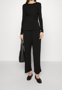 Czarna bluzka z długim rękawem z skręconym designem w talii, w połączeniu z czarnymi spodniami o szerokich nogawkach i płaskimi czarnymi butami. Towarzyszy jej czarna torba.