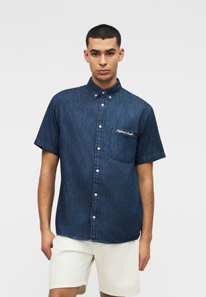 Homme portant une chemise en denim foncé à manches courtes boutonnée avec une poche sur la poitrine et un short blanc, sur un fond uni.