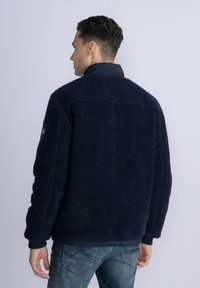 Navy blue fleece jacket with a textured surface, high collar, and ribbed cuffs. Features a contrasting lower fabric and a small logo patch on the sleeve.