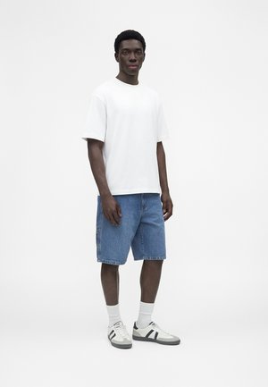 Mees kannab lihtsat valget t-särki, siniseid teksapükse, valgeid sääresokke ja valgeid tossusid mustade triipudega, seisab valge tausta ees.