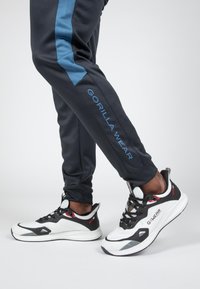 Zwarte en blauwe sportbroek met de tekst "GORILLA WEAR", gecombineerd met zwart-witte sneakers met rode accenten en een gestroomlijnd ontwerp.