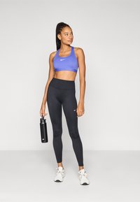 Μπλε αθλητικό σουτιέν με λευκό λογότυπο, συνδυασμένο με μαύρο κολάν ψηλόμεσο. Το άτομο κρατά ένα μαύρο μπουκάλι νερό και φοράει λευκά αθλητικά παπούτσια.