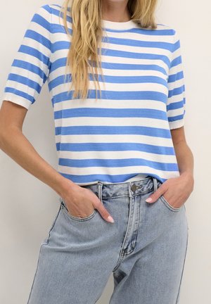 Vrouw draagt een blauw-wit horizontaal gestreept shirt met korte mouwen, ingestopt in een lichtblauwe high-waisted jeans, met haar handen in de zakken.