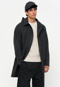 Mustamõõtmetega veekindel jakk terava kraega, nupudega ja pika siluetiga, kantud heledat beeži kudumist-sweatshirt'i ja tumedate pükstega.