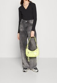 Svart långärmad topp, gråvida byxor med vida ben, håller en skinande neongul handväska med dekorationer och dragkedjedetalj.