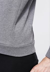 Primer plano de una persona que lleva una sudadera de manga larga gris y pantalones negros, con la mano derecha apoyada cerca de la cadera.