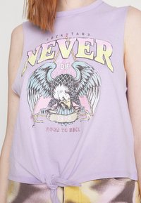 Hihaton laventelinvärinen crop top, jossa on keskeinen kotka grafiikka, rohkea teksti ja solmittu helma. Suunnitelmassa on värikkäitä yksityiskohtia ja vintage-tyyli.