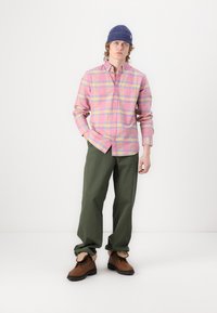 Flanellskjorte i rosa med flerfarget rutemønster, kombinert med grønne bukser og brune støvler. Modellen har på seg en blå strikket lue.