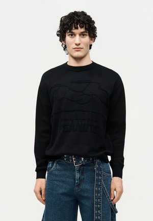 Ung mand med krøllet hår iført en sort Peanuts-trøje og blå jeans med et sort bælte med nitter, stående mod en ensfarvet baggrund.