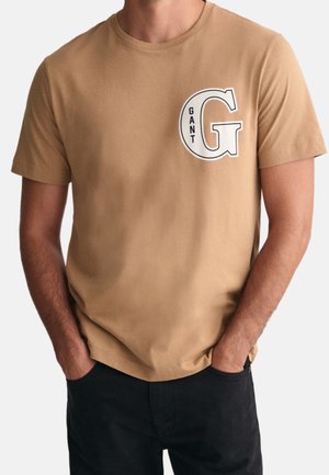 Homme portant un t-shirt beige à manches courtes avec une grande lettre blanche "G" et le mot "GANT" sur le côté gauche de la poitrine, les mains dans les poches d’un pantalon noir.